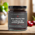 A jar of Sour Cherry Jam from Top Shelf Preserves. The jar is filled with a red jam and has a black lid. The label is black with white text, including the product name, "Handmade Fait à la Main," and instructions to keep refrigerated after opening.