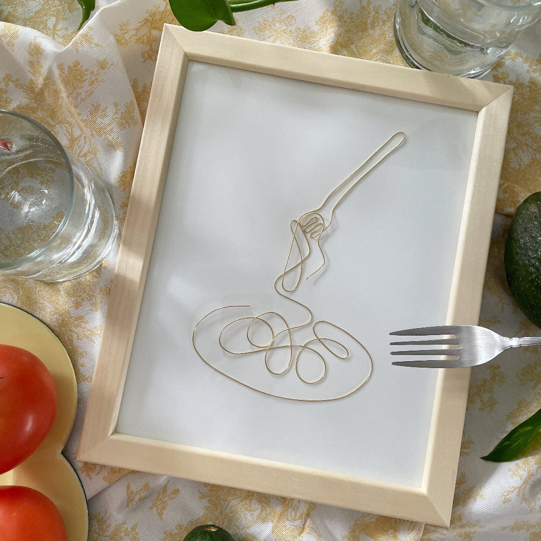 Framed artwork of a fork and spoon on a table with fruits and glasses.