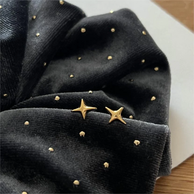A pair of sparkle stud earrings made with 18k gold plating, featuring a minimalist four-pointed star design, displayed on a dark velvet fabric with small gold dots. These celestial earrings are available in Ottawa, Ontario, Canada.