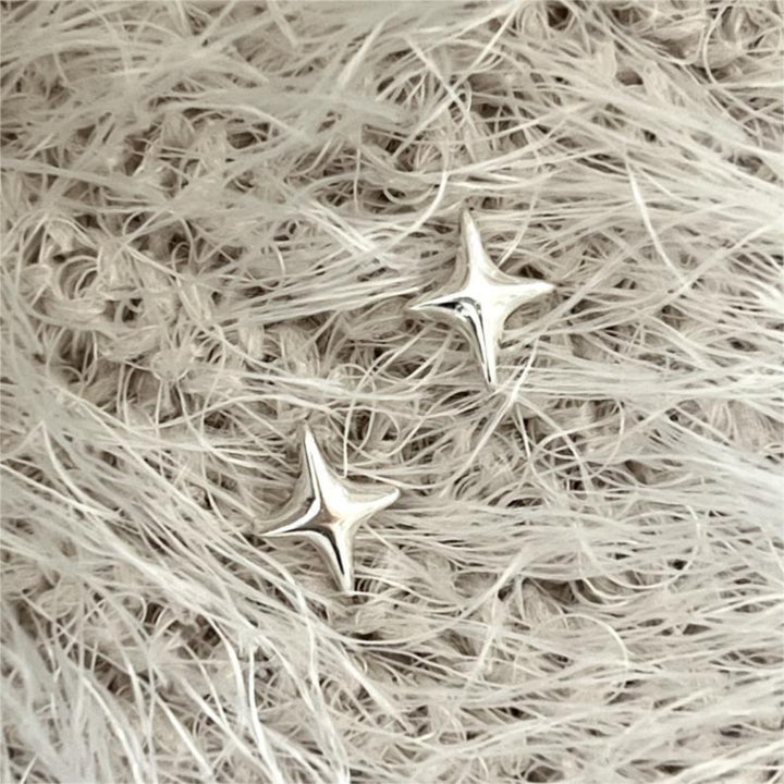 A pair of silver sparkle stud earrings, each featuring a minimalist four-pointed star or starfish-like design, displayed on a textured, light-colored surface. These celestial earrings are available in Ottawa, Ontario, Canada.