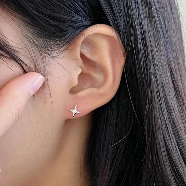 A pair of silver sparkle stud earrings, each featuring a minimalist four-pointed star or starfish-like design, displayed on a textured, light-colored surface. These celestial earrings are available in Ottawa, Ontario, Canada.