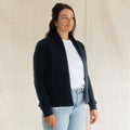 A woman wearing a dark navy blue open-front knit cardigan over a white t-shirt and light-wash blue jeans, standing against a neutral beige fabric backdrop.