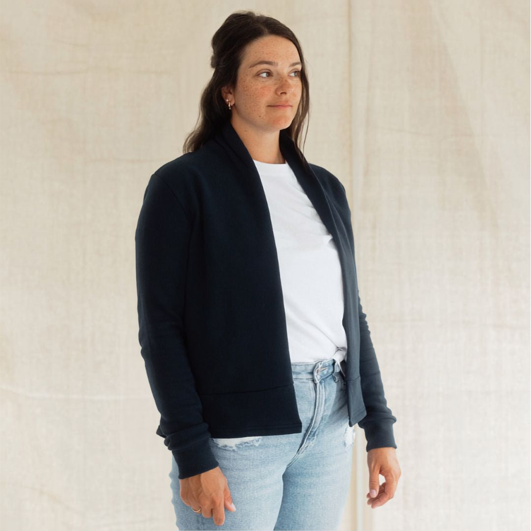 A woman wearing a dark navy blue open-front knit cardigan over a white t-shirt and light-wash blue jeans, standing against a neutral beige fabric backdrop.