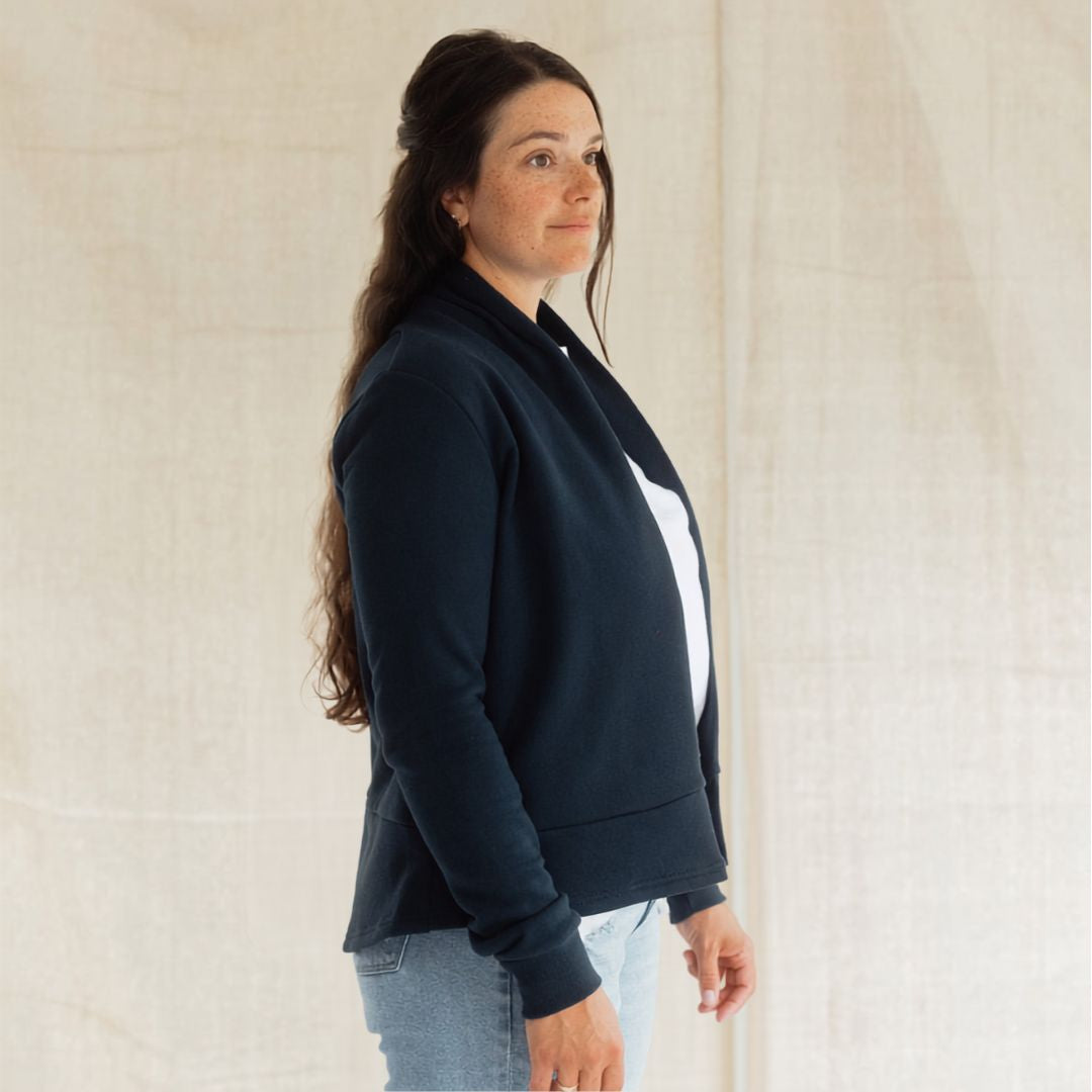A woman wearing a dark navy blue zip-up jacket with a white panel on the front, paired with light blue jeans, standing against a neutral cream-colored fabric backdrop.