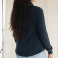 A woman with long dark hair tied back is shown from the back wearing a dark navy long-sleeve quarter-zip fleece jacket and light blue jeans, standing against a light beige fabric backdrop.