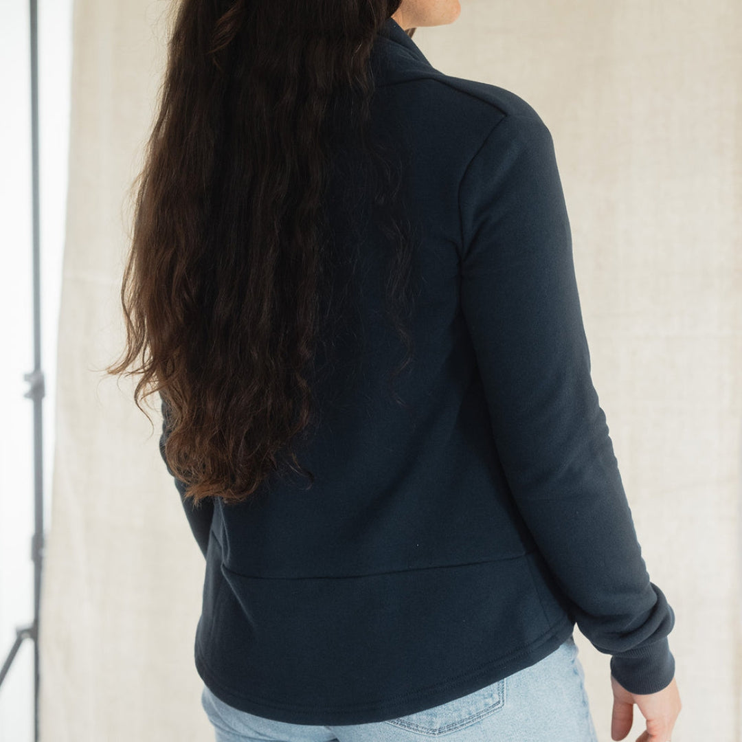 A woman with long dark hair tied back is shown from the back wearing a dark navy long-sleeve quarter-zip fleece jacket and light blue jeans, standing against a light beige fabric backdrop.