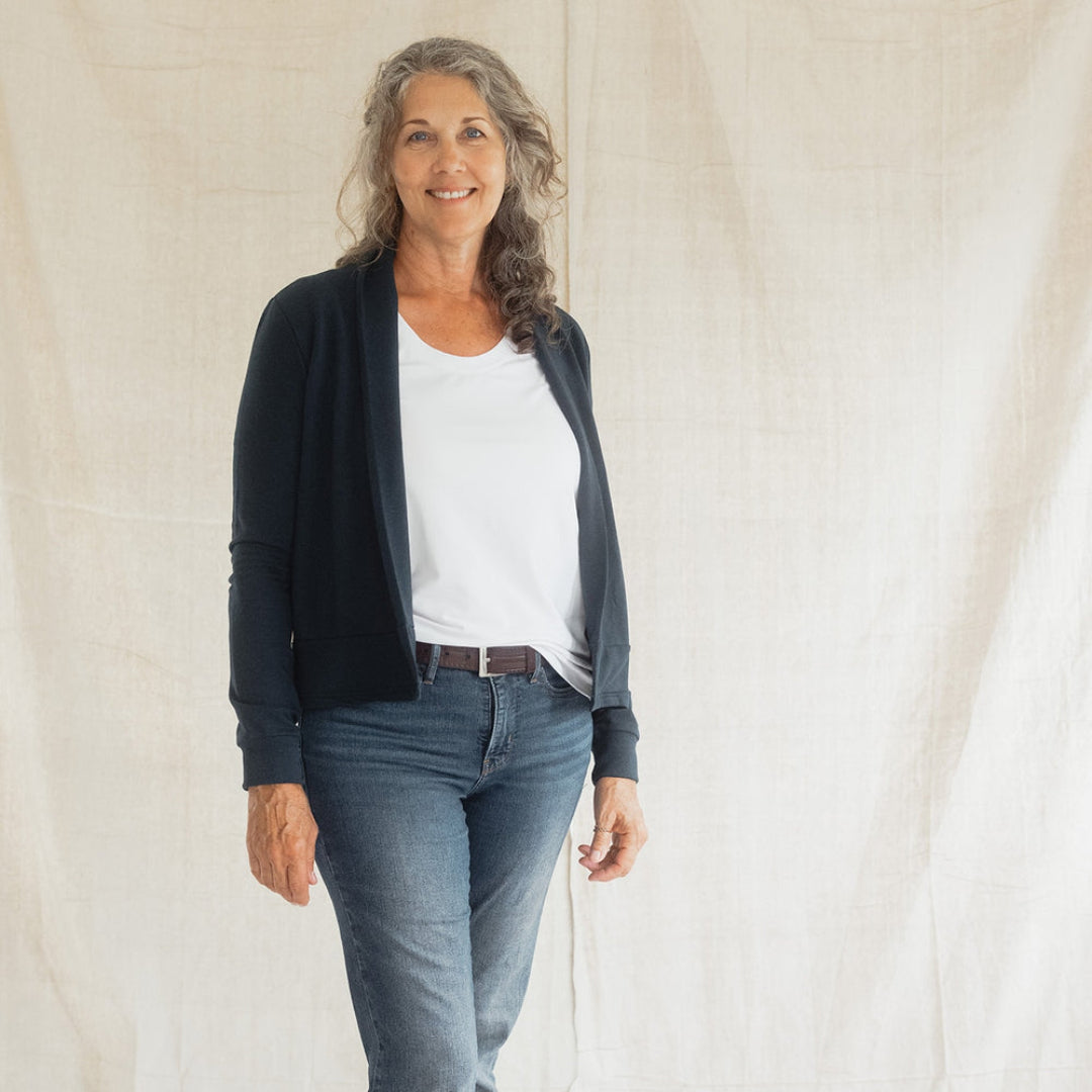 A woman stands barefoot against a draped off-white fabric backdrop, wearing a dark navy cardigan over a white top, blue jeans, and a brown belt.