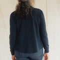 A woman viewed from behind wearing a dark navy blue long-sleeve knit top and blue jeans, standing against a light beige fabric backdrop.
