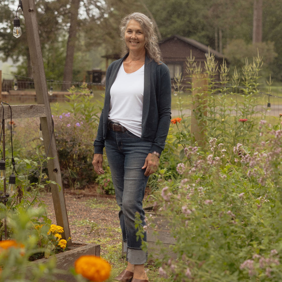A smiling woman with gray hair stands in a garden path wearing a white t-shirt, dark navy cardigan, blue jeans, and brown sandals. The background features lush green foliage, a wooden structure, and orange flowers in the foreground.