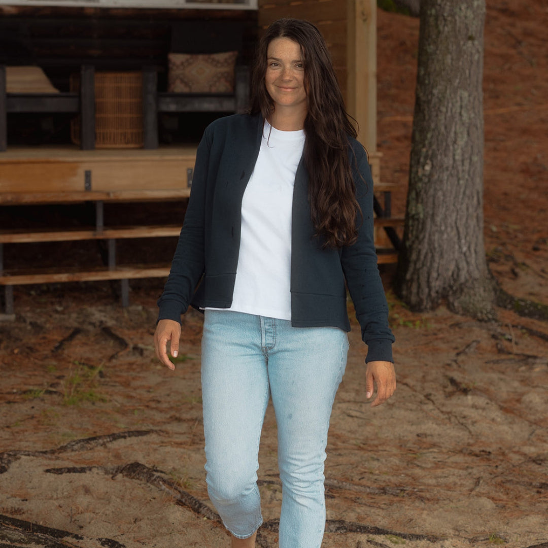 A woman walking barefoot on a dirt path in front of a cabin, wearing a dark navy blue zip-up cardigan over a white t-shirt and light-wash blue jeans.