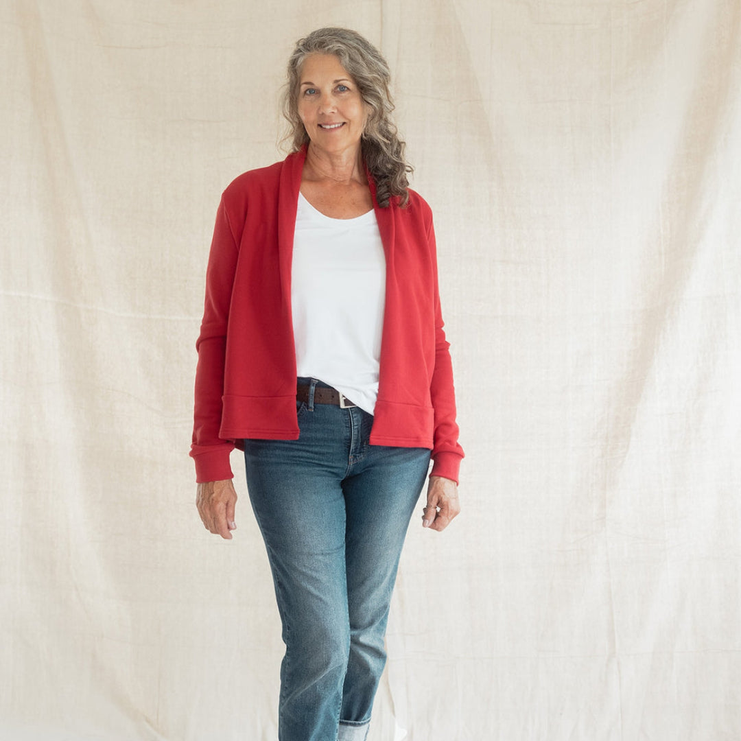 A woman stands barefoot against a draped off-white fabric backdrop, wearing a bright red open-front cardigan over a white top with blue jeans.