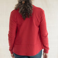 A woman viewed from behind wearing a red long-sleeve top and blue jeans, standing against a light beige fabric backdrop.