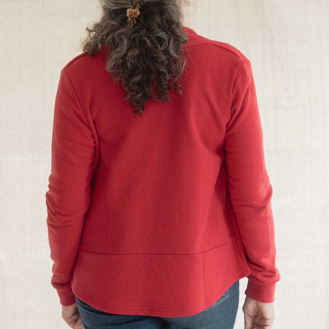 A woman viewed from behind wearing a red long-sleeve top and blue jeans, standing against a light beige fabric backdrop.