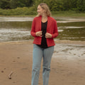 A woman stands on a sandy shore with water and trees in the background, wearing a red open-front jacket over a black top with light blue jeans.