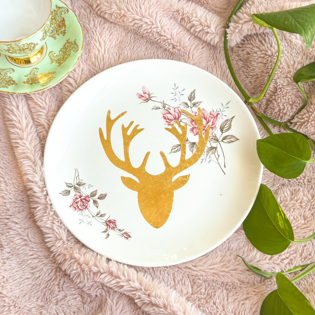 Decorative plate with gold deer head and floral design on a soft pink surface with leaves.