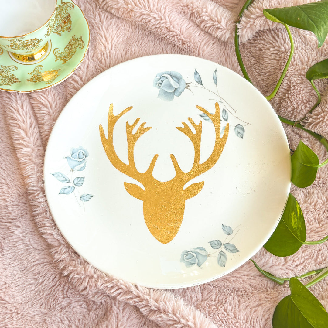 Decorative plate with gold deer head design on a pink textured surface with green leaves.