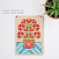 A cross stitch kit on a wooden board with a colorful floral design, styled on a white background next to a potted succulent. Text on the image reads 'cross stitch kit with wood shape'.