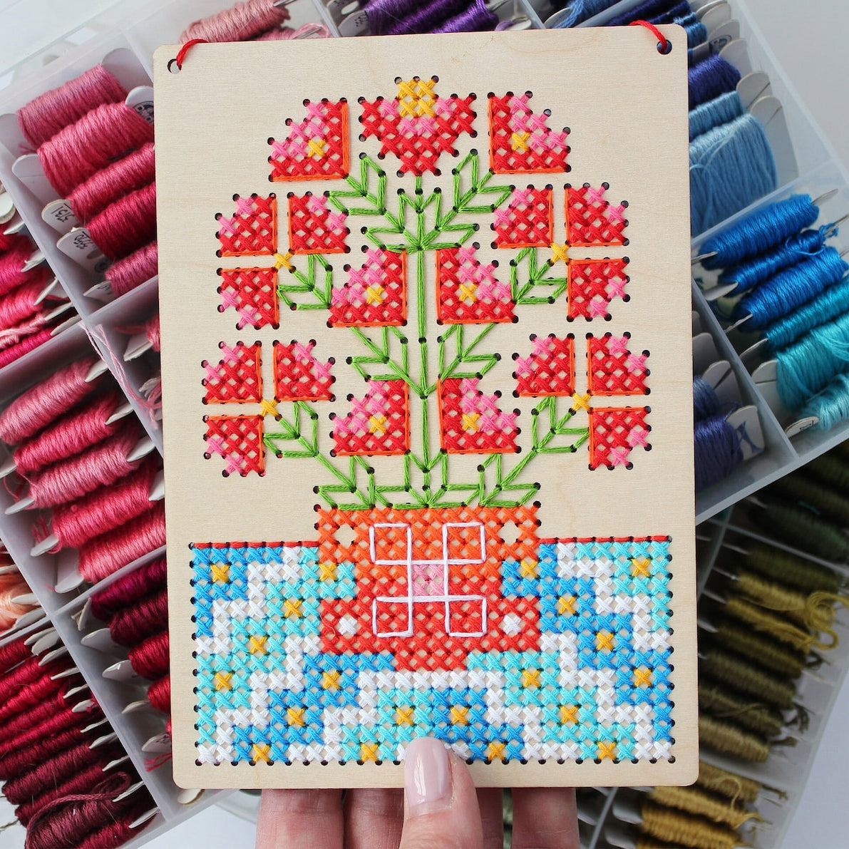 A wooden cross-stitch sampler board featuring a colorful floral design, held by a hand in front of a large organizer filled with spools of thread in various colors.
