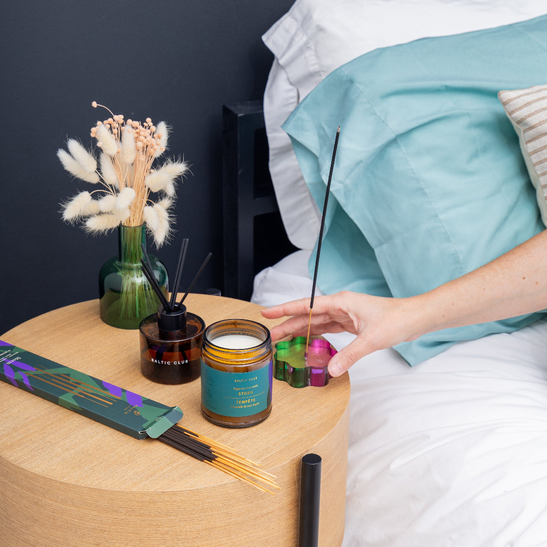 Person holding incense sticks next to a candle and diffuser on a wooden table.