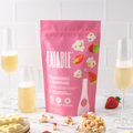 Pink package of 'Eatable' strawberries and champagne popcorn on a white surface with glasses of champagne and strawberries.