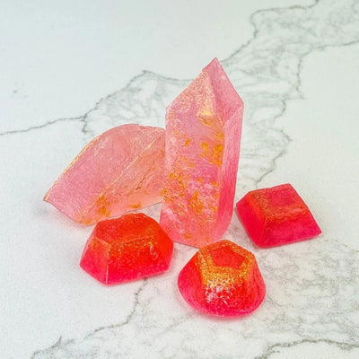 Several pink, crystal-shaped candies arranged on a white marble surface. Some candies are a pale pink, while others are a darker shade with gold accents. The candies have a crystal-like appearance with a slightly translucent quality.