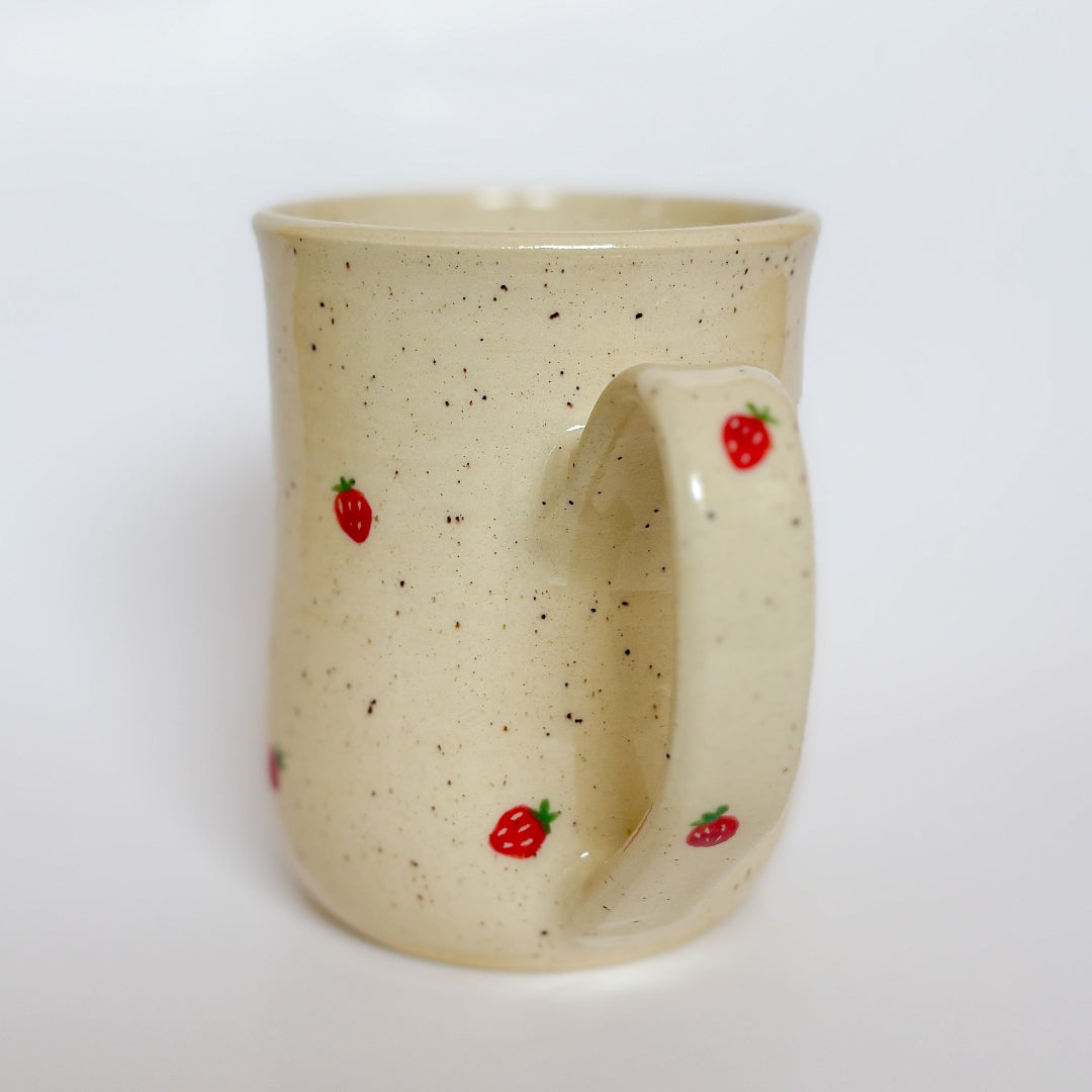 A handmade ceramic mug by Valerie Daoust Ceramics, featuring hand-painted red strawberries on a speckled cream background, displayed on a light gray background. The mug has a large, rounded handle, a speckled texture, and is made in Mississippi Mills, Ontario, Canada.
