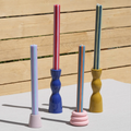 Four decorative taper candle holders in pastel colors, each holding a striped candle, arranged on a light surface against a wooden slat wall.