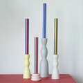 Four decorative taper candle holders in pastel and textured finishes, each holding a tall candle, displayed on a red cabinet against a light gray wall.