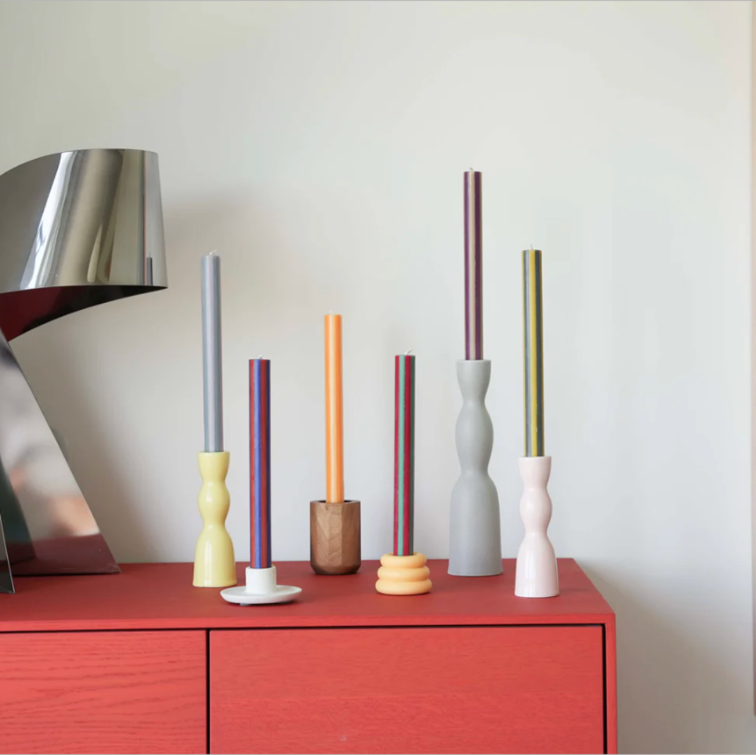 A set of six minimalist ceramic candlesticks in various colors and shapes, displayed on a red wooden cabinet next to a modern metallic table lamp against a white wall.