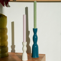 Two matte ceramic candle holders on a wooden shelf against a white wall. One holder is white with a light pink candle, and the other is a dark teal-blue with a sage green candle.