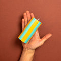 A hand holding a striped pillar candle with alternating yellow and light blue-green stripes against a solid terracotta brown background.
