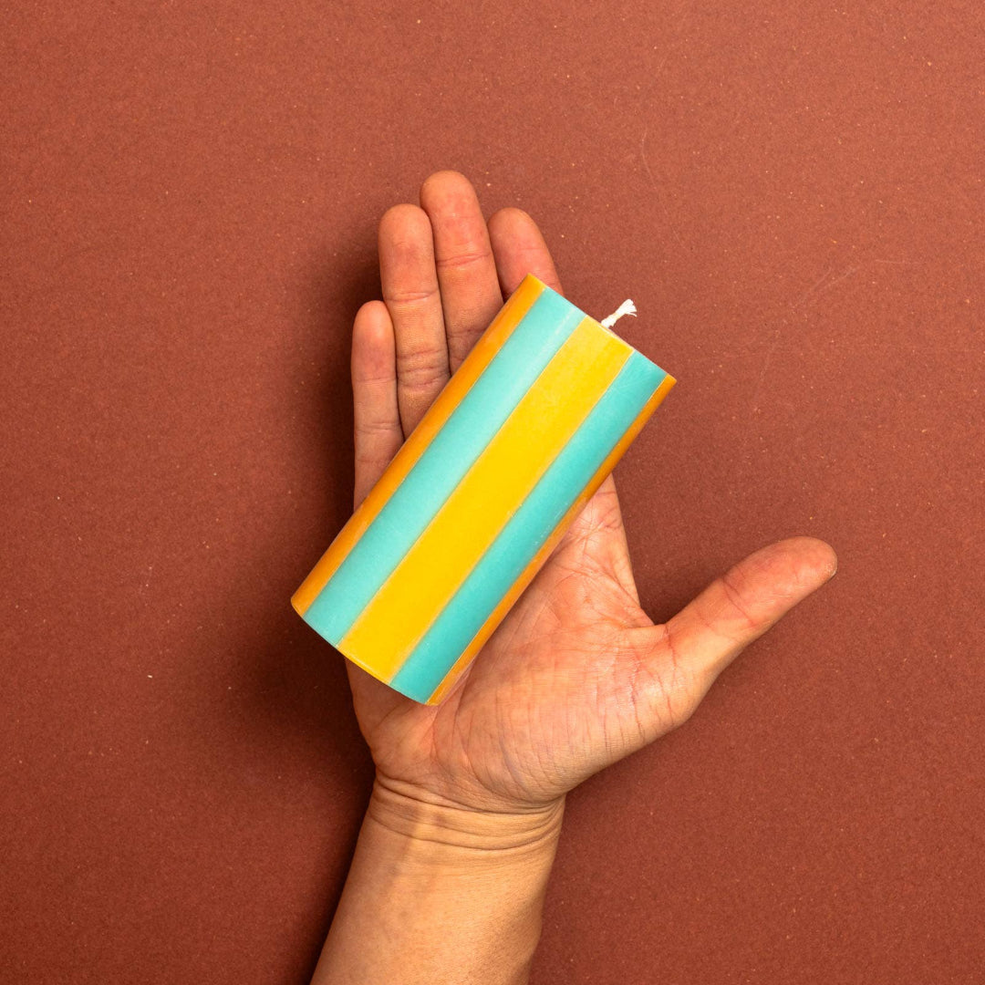 A hand holding a striped pillar candle with alternating yellow and light blue-green stripes against a solid terracotta brown background.