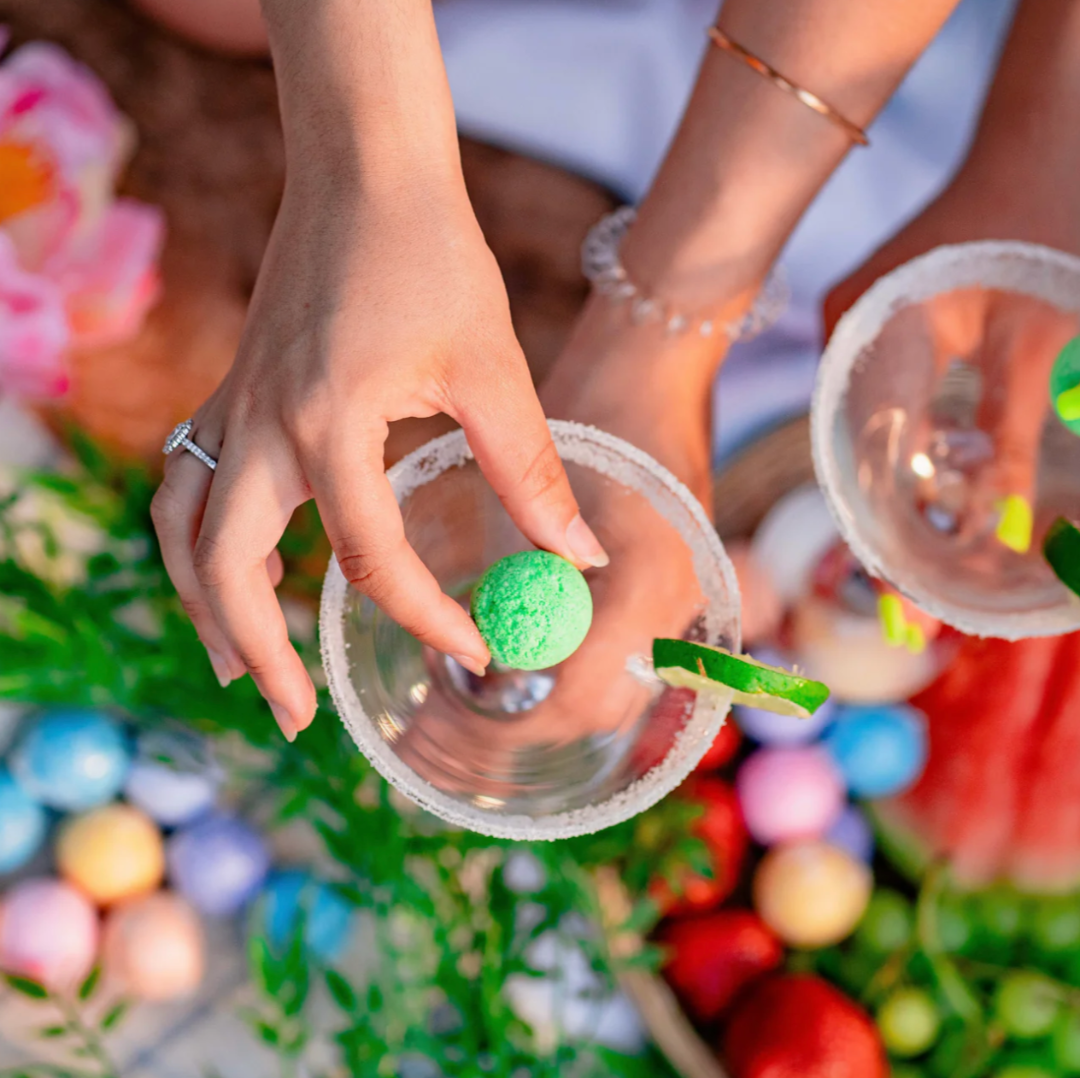 A Summer Cocktail Bomb Variety Pack, featuring colorful, fruit-flavored drink mixers in a white box, with a hand picking up a yellow cocktail bomb. The image includes strawberries, pineapples, and limes against a bright blue background. Available in Ottawa, Ontario, Canada.
