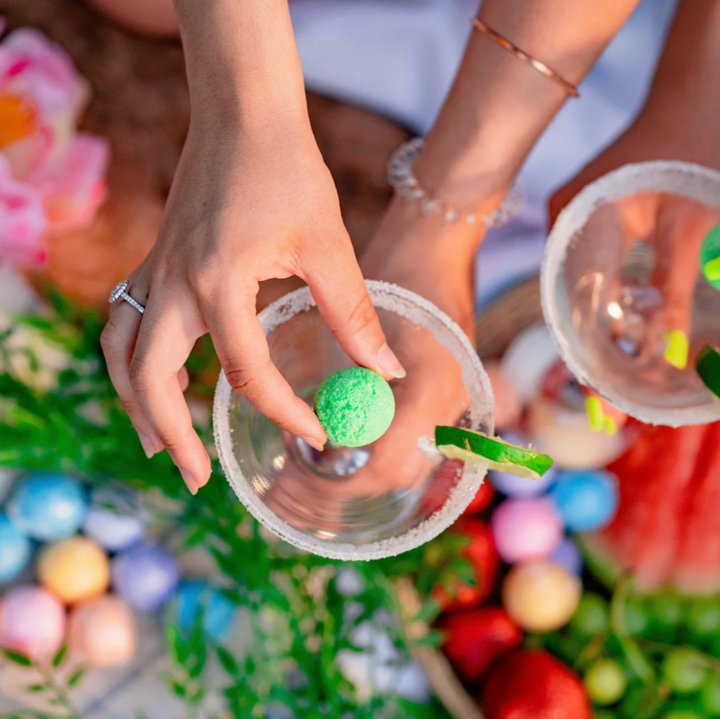 A Summer Cocktail Bomb Variety Pack, featuring colorful, fruit-flavored drink mixers in a white box, with a hand picking up a yellow cocktail bomb. The image includes strawberries, pineapples, and limes against a bright blue background. Available in Ottawa, Ontario, Canada.