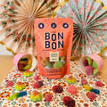A stand-up pouch of Bon Bon candy stands on a floral tablecloth with loose gummy candies scattered around it. Two pink cups tipped over on the left and right show more gummies.