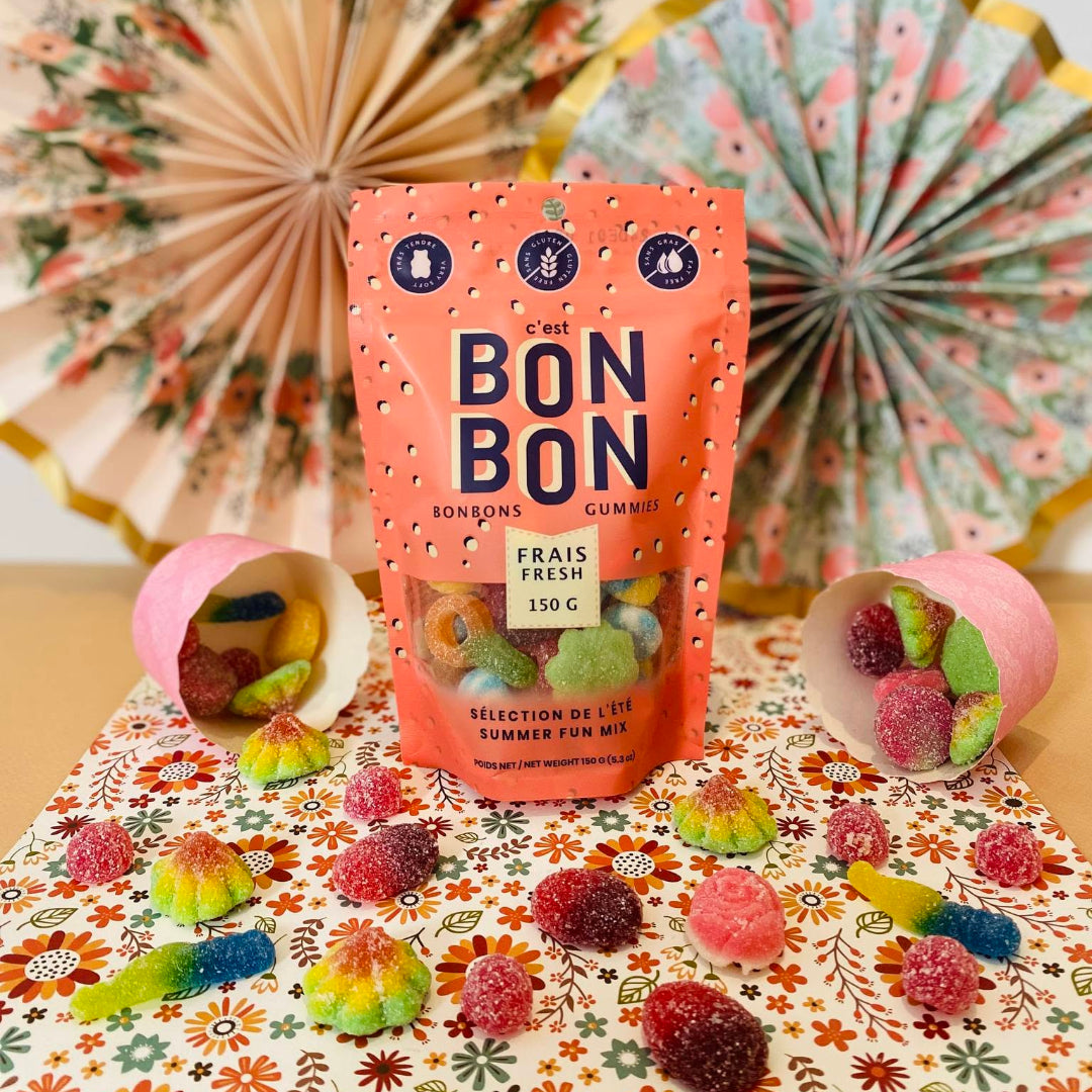 A stand-up pouch of Bon Bon candy stands on a floral tablecloth with loose gummy candies scattered around it. Two pink cups tipped over on the left and right show more gummies.