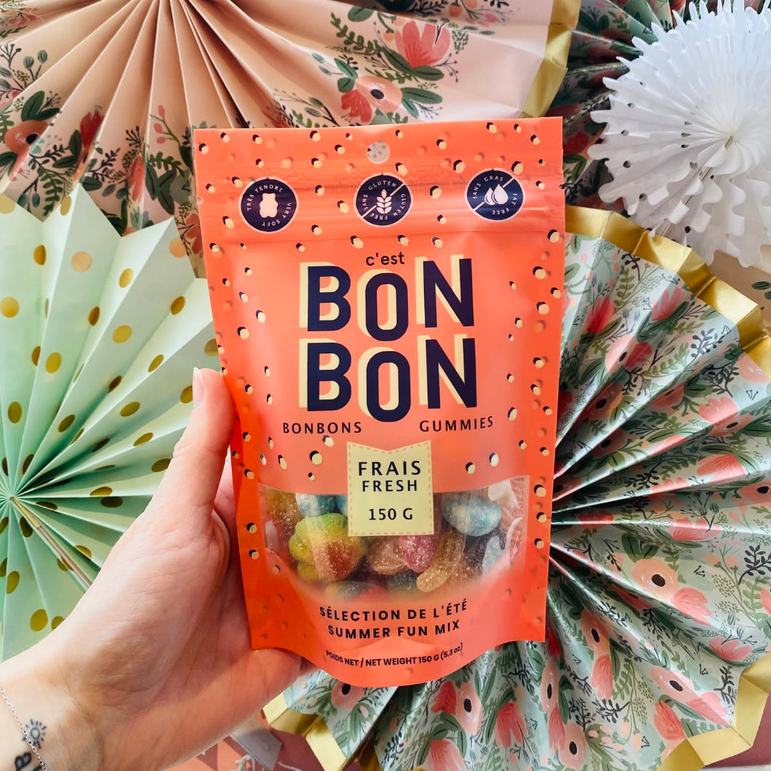 A hand holds a bag of c'est BON BON Sconeons Gummies against a background of decorative paper fans in floral and polka-dot patterns.