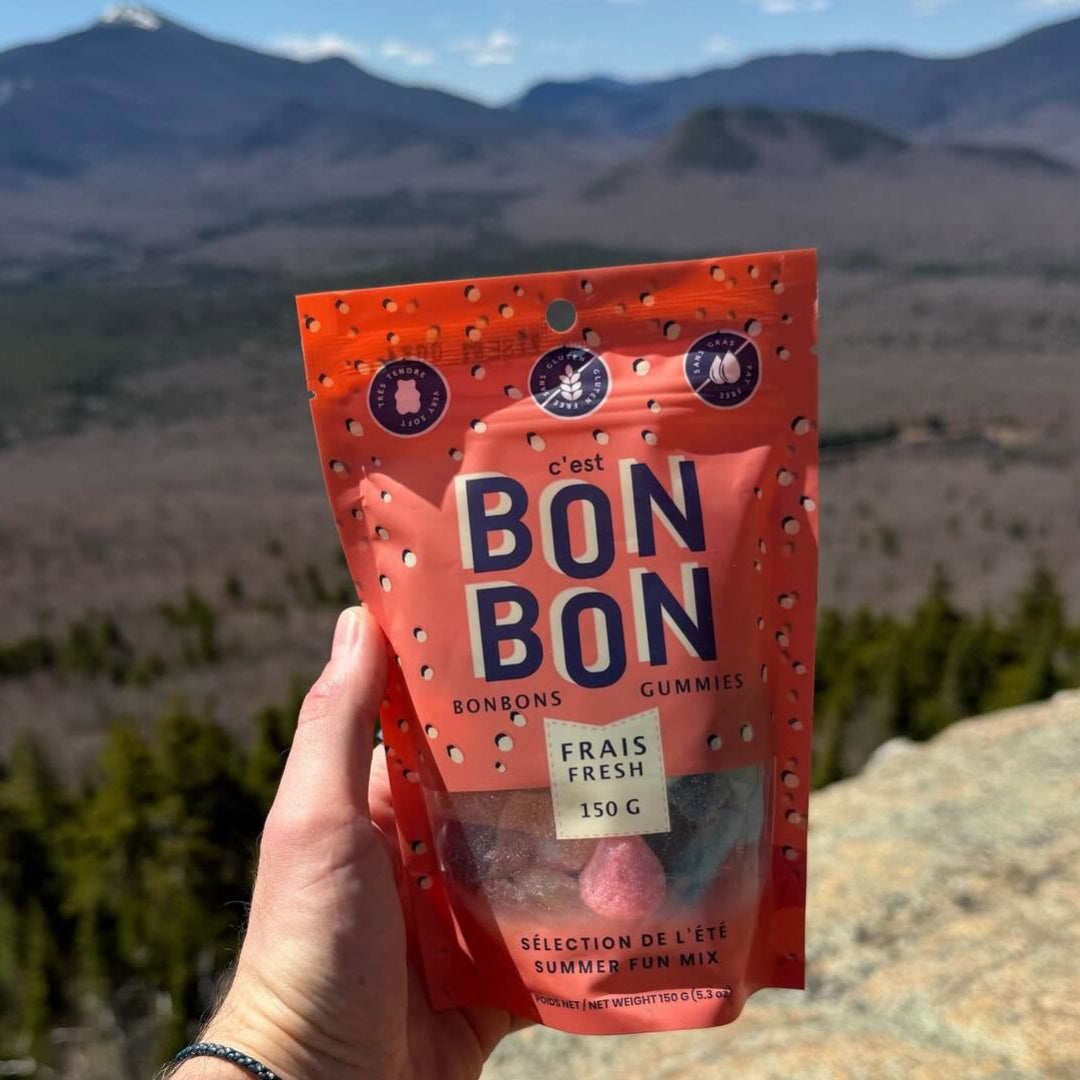 A hand holds a red bag of Bon Bon gummies in front of a mountain landscape. The packaging is bright red with dark blue lettering and small white dots, with the brand name 'Bon Bon' clearly visible.