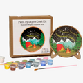 Paint by layers craft kit with a completed painting of a tent and campfire, surrounded by paint containers and a brush.