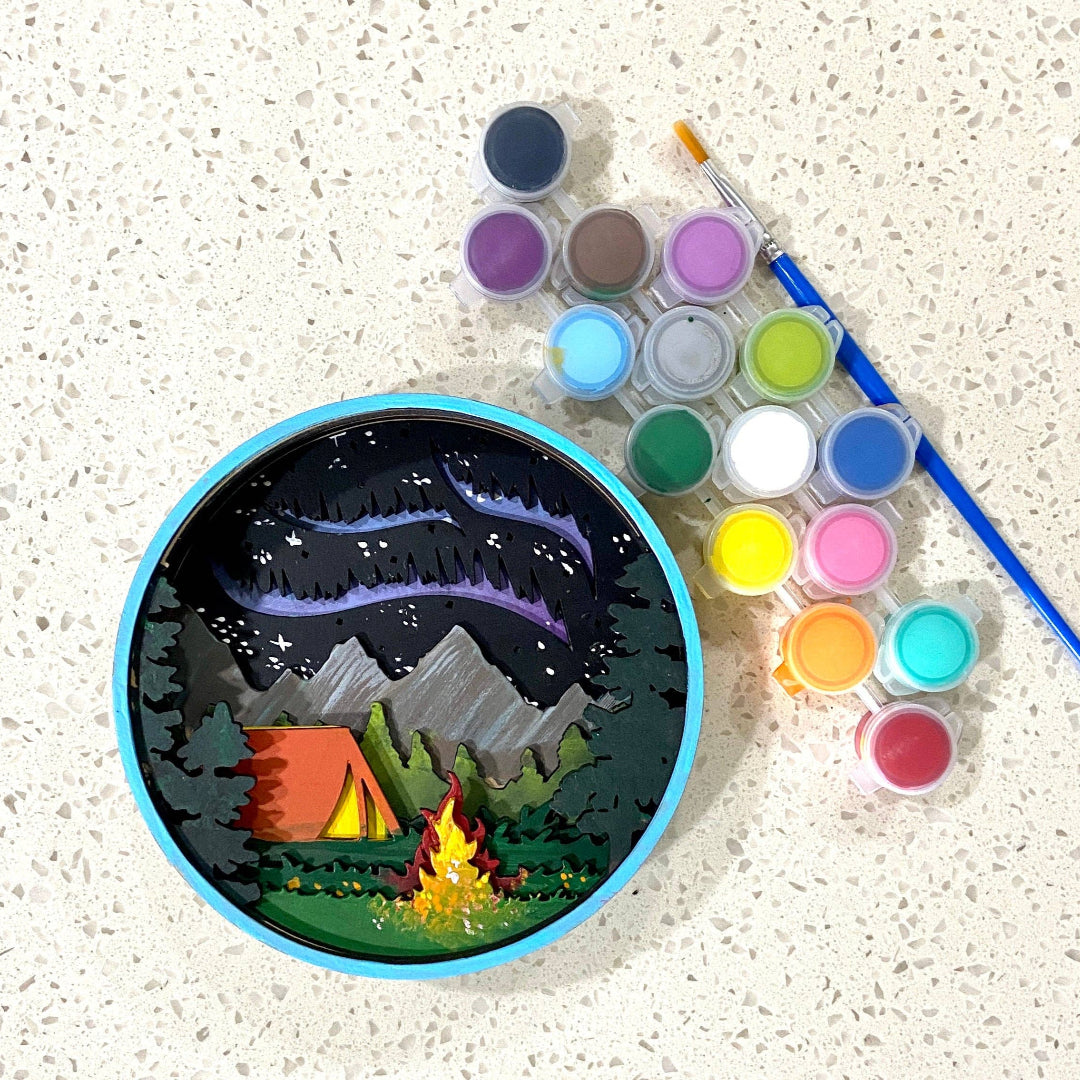 Painting of a camping scene with watercolor paints and brushes on a textured surface