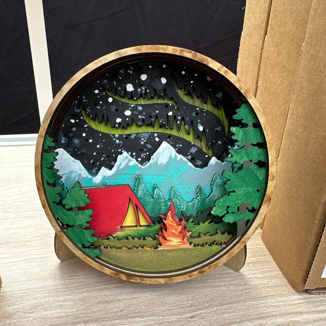Round wooden art piece with a camping scene including a red tent, campfire, and mountains.