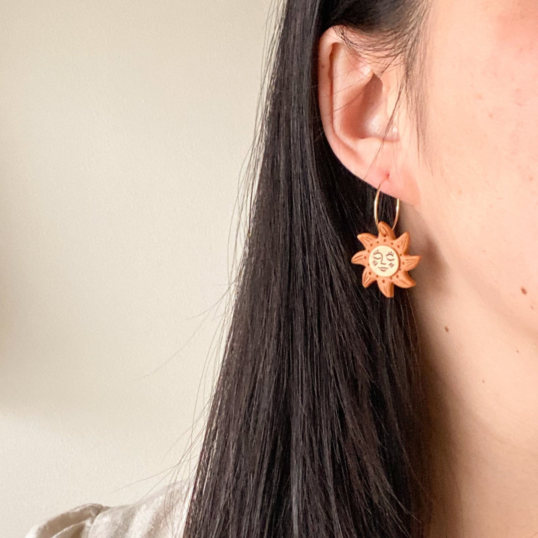 Sun-shaped earrings worn by a person with long dark hair against a neutral background