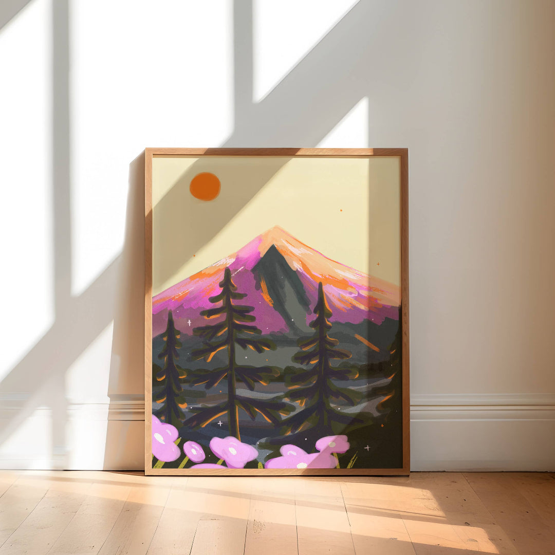A framed art print of a stylized mountain landscape with pine trees and pink flowers, leaning against a white wall on a light wood floor in a sunlit room.