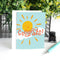 A congratulations card featuring a smiling sun with rays shining on clouds. The text 
