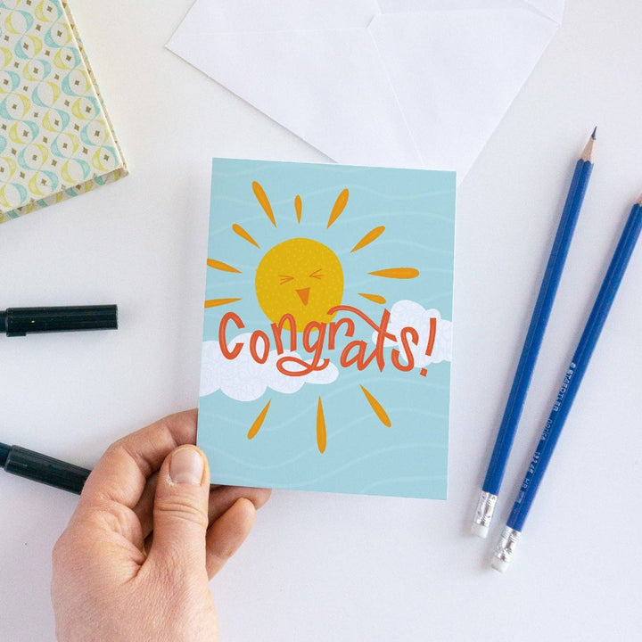 A congratulations card featuring a smiling sun with rays shining on clouds. The text 