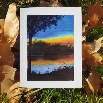 A watercolor greeting card of a serene lakeside scene at sunset. A dark silhouette of a tree stands tall in the foreground, with a vibrant orange and yellow sky reflected in the calm water.