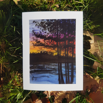A watercolor greeting card of a sunset over a still lake. The sky is ablaze with vibrant orange, yellow, and purple hues, reflected in the calm water. Silhouettes of tall trees stand in the foreground, creating a dramatic contrast against the colorful sky.