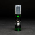 A dark green glass bottle with a black pump dispenser. The bottle is labeled "BUSH" and "SWEET BEARD OIL." The background is black.