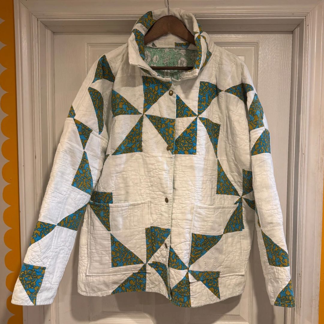 A vintage quilted jacket with a geometric triangle pattern in teal, blue, and yellow on an off-white background, hanging on a wooden hanger against a white paneled door.