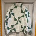 A quilted jacket with a high mock neck and hood, featuring a white base with geometric triangles patterned in teal and yellow, hanging on a wooden hanger in front of a white paneled door.
